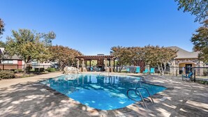 Ein rechteckiger Pool mit einer Pergola und Liegestühlen, umgeben von Bäumen und einem Zaun.