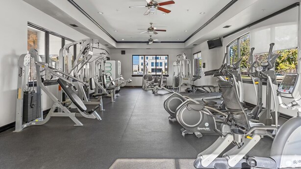 Ein Fitnessstudio mit verschiedenen Trainingsgeräten, Deckenventilatoren und großen Fenstern.
