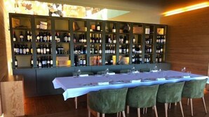 A dining area with a long table set for six, surrounded by green upholstered chairs. Behind the table, there is a wall of wine bottles on shelves. - Monverde - Wine Experience Hotel - by Unlock Hotels (Borba de Godim)