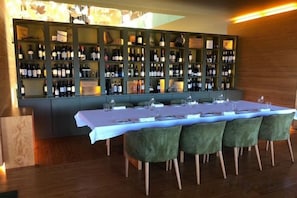 Una larga mesa con mantel blanco, sillas tapizadas en verde y una estantería para vinos al fondo.