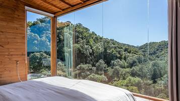 Een slaapkamer met een groot raam dat uitkijkt over een bebost glooiend landschap.