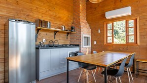A compact kitchen with a refrigerator, microwave, stove, and oven. There is a wooden dining table with chairs and a window with a view of greenery. - Cabana vista Serra Catarinense VST023 (Anitápolis)
