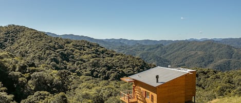 A wooden cabin with a balcony, surrounded by forested hills.