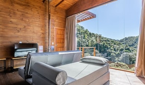 Una cabana de fusta amb un sofà i una televisió sobre una taula de fusta, una finestra gran amb vista a una collada boscosa.