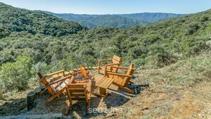 A wooden table and chairs set up outdoors with a fire pit in the center, surrounded by a mountainous landscape. - Cabana vista Serra Catarinense VST023 (Anitápolis)