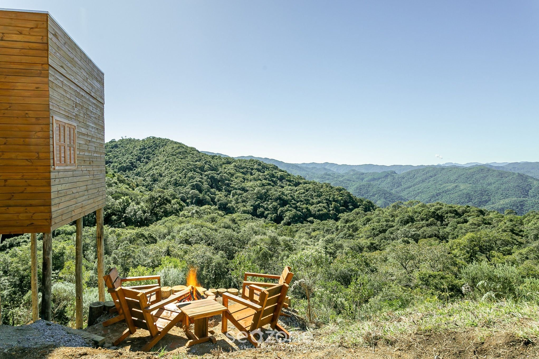 Een houten hutje met een vuurplaats en stoelen met uitzicht op een bebost berglandschap.