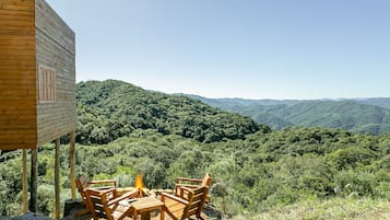 A wooden cabin with a fire pit and chairs overlooking a forested mountain range.
