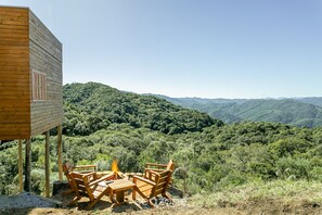 Una cabaña de madera con un hogar a la vista y sillas, con vista a una cadena montañosa boscosa.