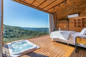 Habitación de cabaña de madera con cama, jacuzzi y un amplio ventanal con vistas a una cadena montañosa boscosa.