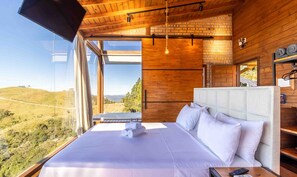 A modern bedroom with a large bed, a view of the countryside, and wooden walls. - Cabana espetacular região serrana VST008 (Anitápolis)