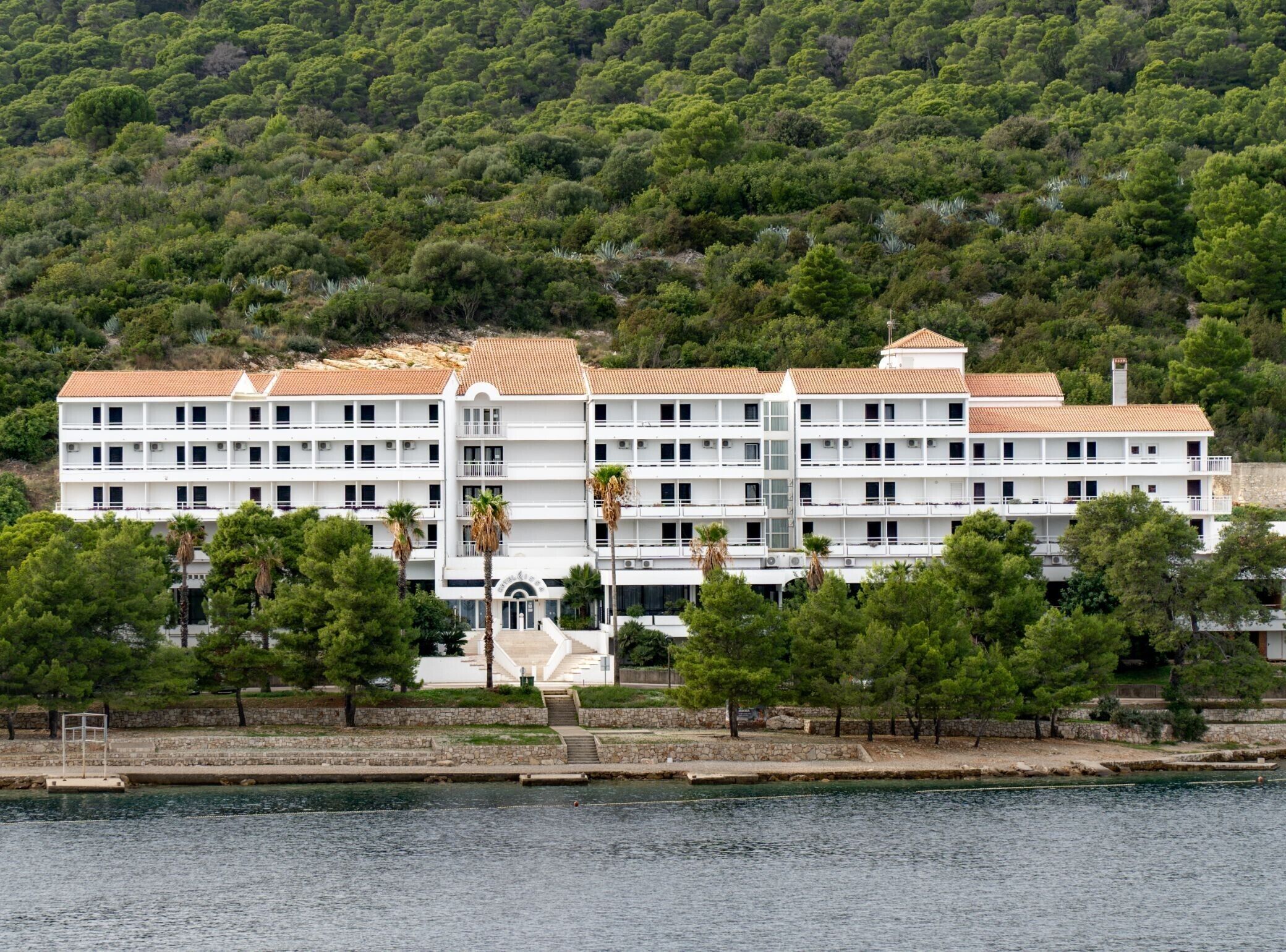 Un hôtel à plusieurs étages, doté d’une façade blanche et d’un toit recouvert de tuiles rouges, situé au bord de l’eau, avec un mur de pierre et des palmiers au premier plan.