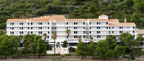 A multi-story hotel with a white facade and red-tiled roof, situated by a waterfront with a stone wall and palm trees in the foreground.