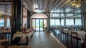 A spacious restaurant with a high ceiling, modern lighting fixtures, and a central archway leading to an outdoor area. Tables are neatly arranged, and the floor is tiled. - Hotel Issa - Double Room Sea View 14 (Vis)