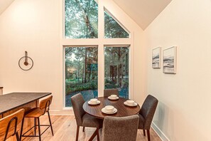 A dining area with a round table, six chairs, and a view of a forest through large windows.