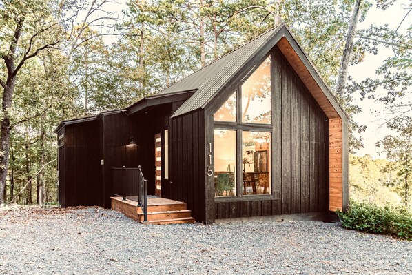 A rustic cabin with a large window, wooden exterior, and a gravel driveway.