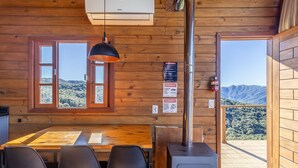 Interior de una cabaña de madera con una mesa de comedor, sillas, una lámpara colgante, una estufa y vistas a las montañas a través de una ventana.