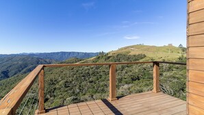Une terrasse en bois, dotée d’une rambarde en métal, offre une vue sur un paysage vallonné avec des montagnes à l’horizon.