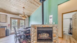 Une cheminée en pierre avec une hotte, un coin repas avec une table et des chaises, et une vue sur l’extérieur par une porte.