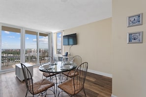 Un coin repas avec une table et des chaises en verre, un téléviseur à écran plat fixé au mur et une vue sur la ville grâce à de grandes fenêtres.