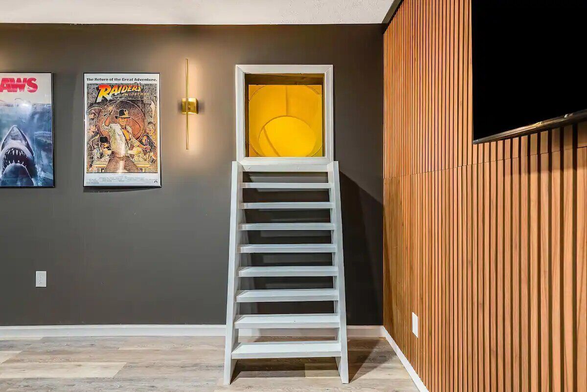 Una escalera con estructura blanca que conduce a un cuarto iluminado con luz amarilla, frente a una pared oscura con dos carteles de películas.