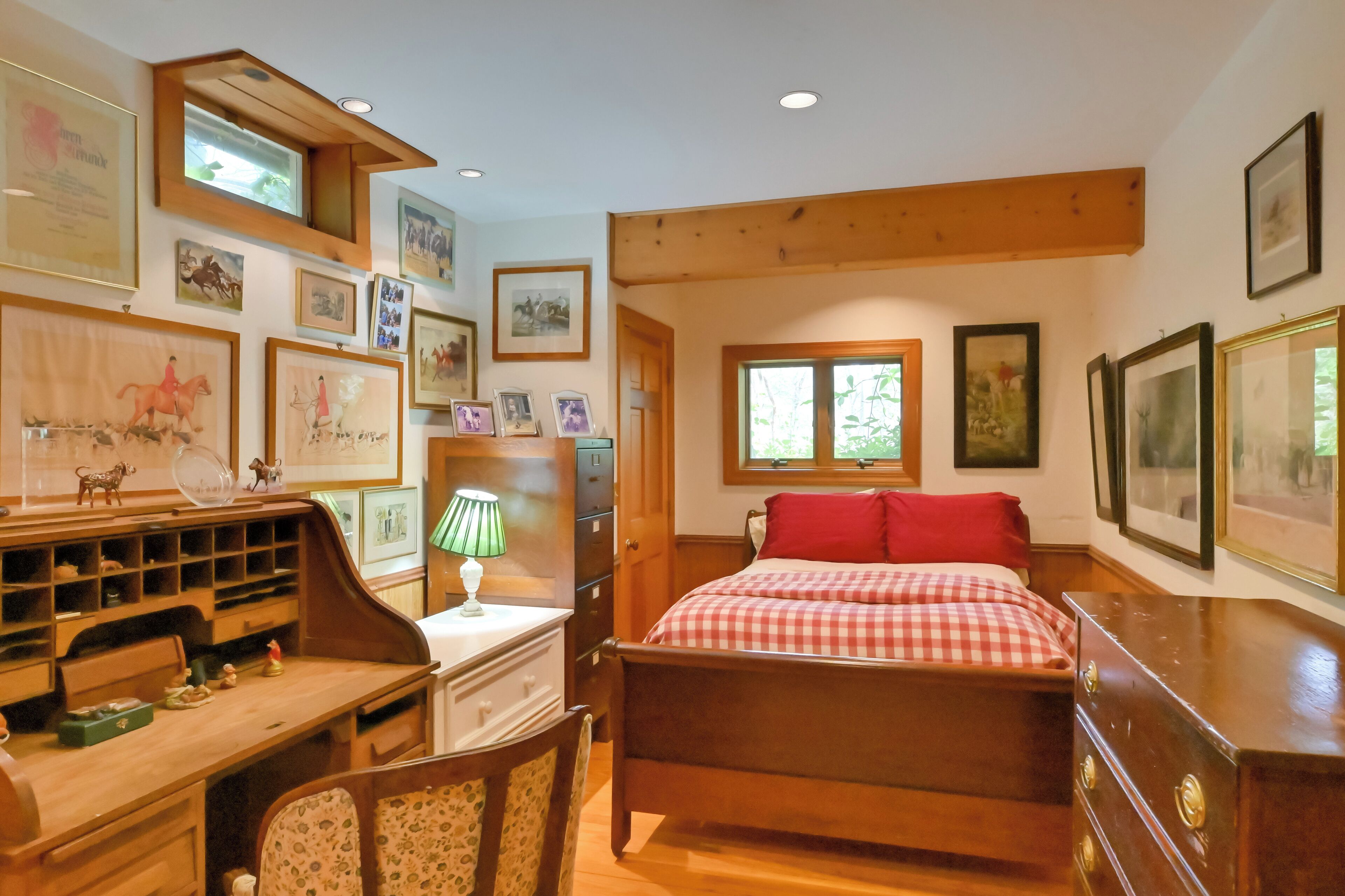 A bedroom with a wooden bed, a desk with a lamp, and various framed pictures on the walls.