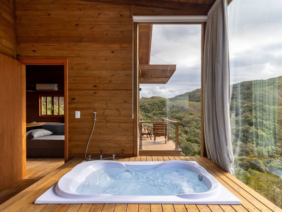A wooden cabin with a hot tub, a bedroom with a bed, and a balcony with a view of the hills.