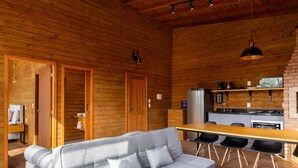 Un acogedor interior de cabaña, con paredes y mobiliario de madera, un sofá, una mesa de comedor con sillas, una zona de cocina con electrodomésticos y una lámpara colgante.