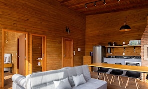 Interior acollidor d'una cabana amb elements de fusta, un sofà, una taula de menjador amb cadires, una zona de cuina amb electrodomèstics i un llum penjant.
