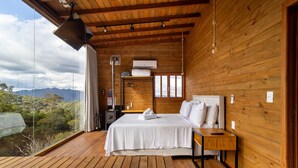 A wooden cabin bedroom with a bed, bedside table, and a view of the outdoors. - Cabana c/ vista e jacuzzi Serra Catarinense VST025 (Anitápolis)