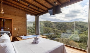 Una habitación con techo de madera, un ventanal con vistas a un paisaje montañoso, una cama con ropa blanca y una pequeña bañera de hidromasaje.