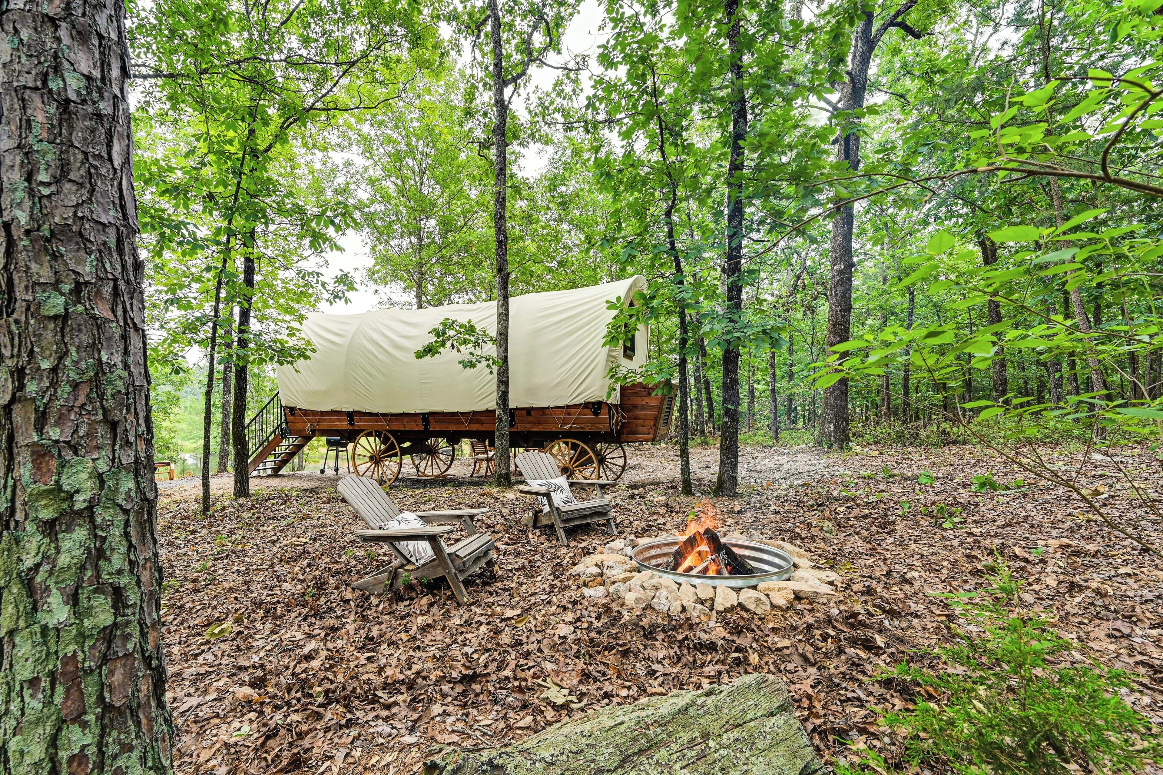 A rustic campsite with a covered wagon, wooden chairs, and a fire pit surrounded by trees.