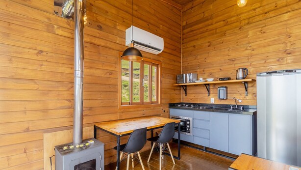 A wooden cabin interior with a kitchen area, a dining table with chairs, a wood stove, and a window with a view of the outdoors. - Cabana incrível com vista VST010 (Anitápolis)