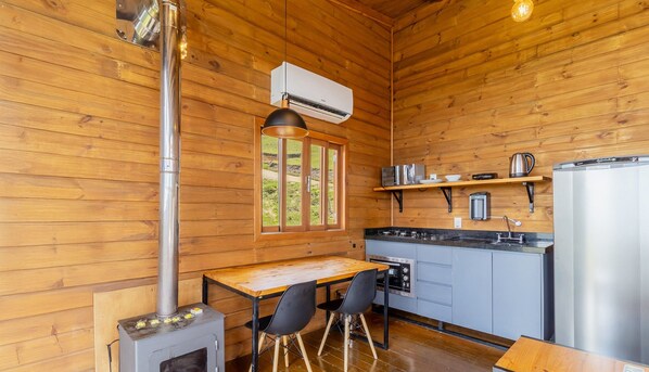 A wooden cabin interior with a kitchen area, a dining table with chairs, a wood stove, and a window with a view of the outdoors.