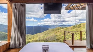 A room with a bed, wooden flooring, a TV, and a view of a mountainous landscape.