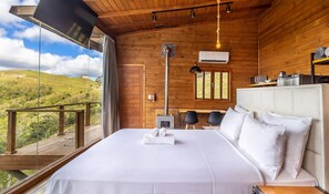 A bedroom with a large bed, a wooden ceiling, a window with a view of the countryside, and a small table with chairs. - Cabana incrível com vista VST010 (Anitápolis)