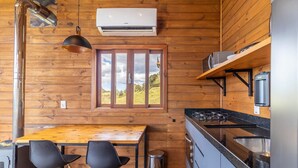 A wooden cabin kitchen with a dining table, two chairs, a window with a view, a stove, and an air conditioning unit. - Cabana incrível com vista VST010 (Anitápolis)