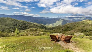 A wooden table and chairs set on a grassy hillside with a view of distant mountains under a partly cloudy sky. - Cabana incrível com vista VST010 (Anitápolis)