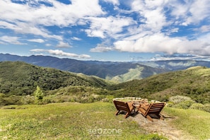 A wooden table and chairs set on a grassy hillside with a view of distant mountains under a partly cloudy sky.