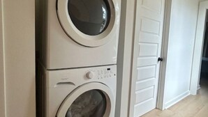 Una lavadora y secadora apiladas en un pasillo con una puerta blanca y piso de madera.