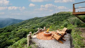 Un foyer circulaire entouré de sièges, lui-même bordé d’une zone recouverte de gravier, le tout se fondant dans un paysage de collines verdoyantes et sous un ciel dégagé.