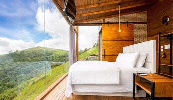 Un dormitorio con una cama grande, suelo de madera, vista a un paisaje verde y una pared de ladrillo.