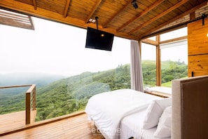 A bedroom with a large bed, a TV mounted on the ceiling, and a view of a forested hillside.