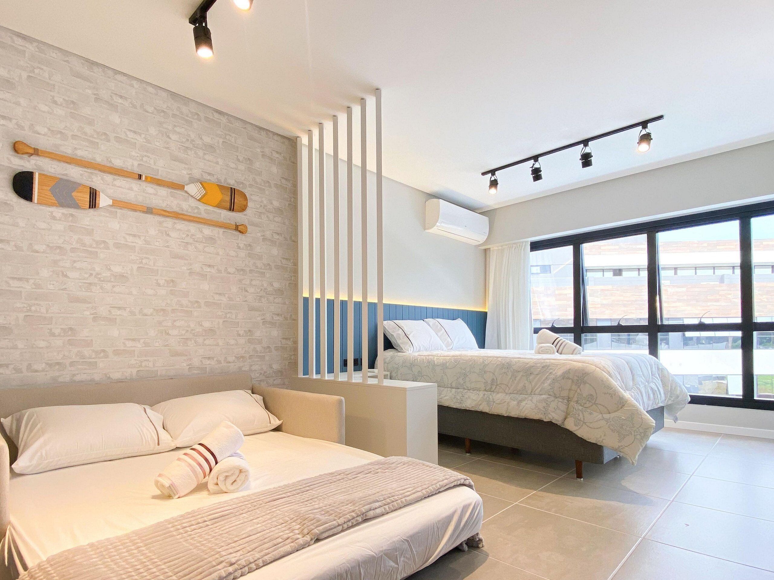 A modern hotel room with a brick wall, a large bed, a smaller bed, a window with a view, and a ceiling light fixture.