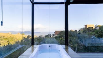 A hot tub in a modern glass-enclosed room with a view of a hilly landscape.