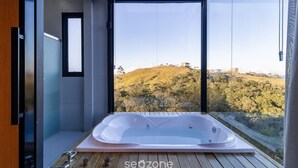 A modern bathroom with a large bathtub, a glass shower enclosure, and a view of a hilly landscape. - Cabana jacuzzi e vista montanhas VST031 (Anitápolis)