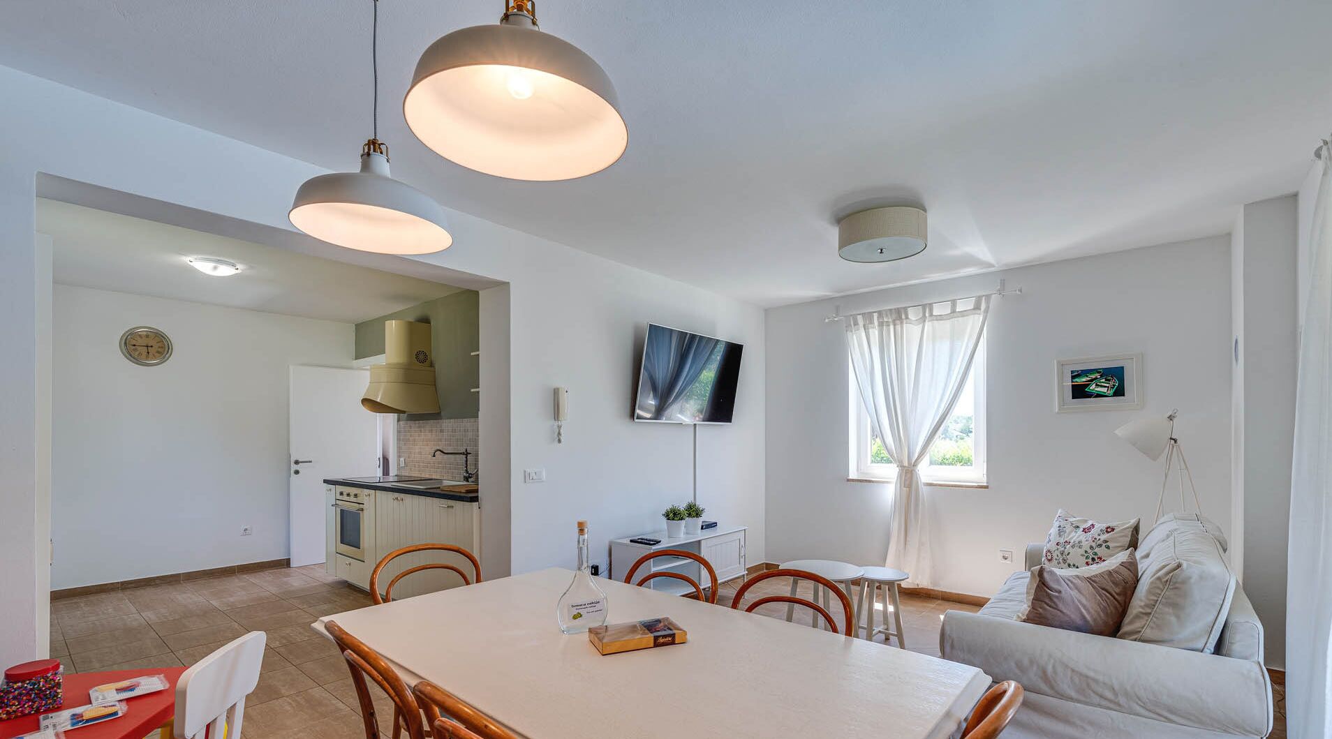 A modern living room with a dining area, a kitchen in the background, and a TV mounted on the wall.