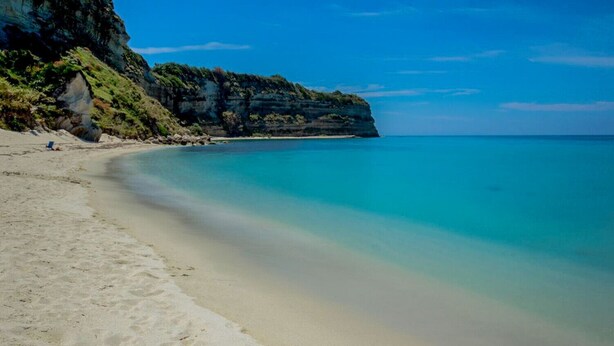 Une plage à l’eau turquoise et limpide, avec un rivage sablonneux et une falaise rocheuse couverte de végétation.