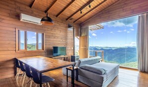 A wooden interior with a dining table, chairs, a sofa, a TV, and a large window offering a mountain view. - Cabana na serra com linda vista VST012 (Anitapolis)