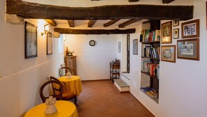 Un couloir étroit aux poutres apparentes, un coin repas avec une table et des chaises jaunes, et une bibliothèque remplie de livres et de photos encadrées.