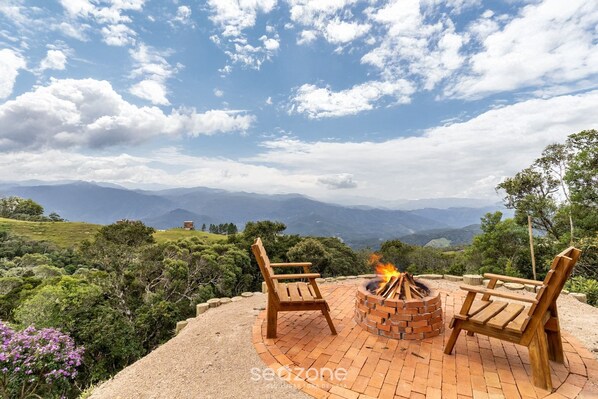 A patio with two wooden chairs and a brick fire pit, overlooking a mountainous landscape.